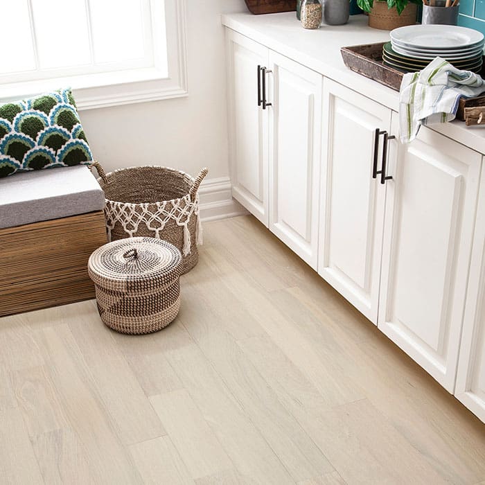 Small kitchen nook area with cushioned bench and white kitchen cabinets. Round, wicker baskets sit on top of light-grained engineered hardwood flooring.