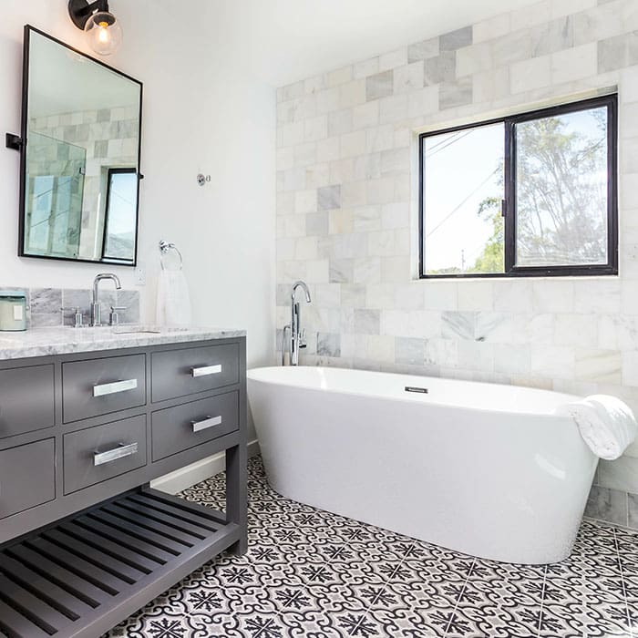 Bathroom with a dark-gray vanity, black iron rectangular mirror, large roman-style white porcelain tub, and holy-leaf patterned black and white flooring tile.