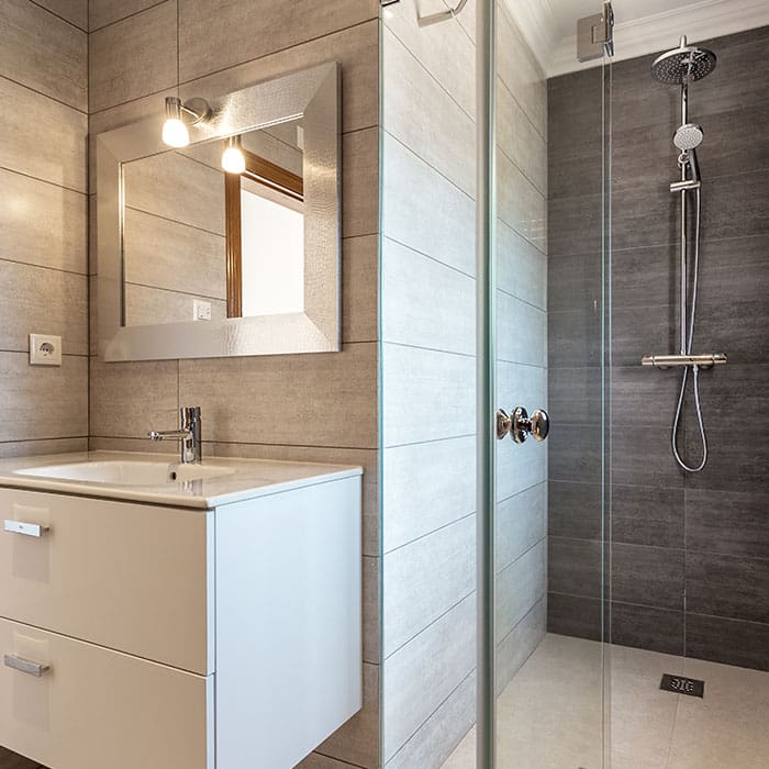 Small bathroom with floor-to-ceiling, medium-textured porcelain tiles. A glass shower wall opens up to a small vanity with silver-framed mirror and modern lighting.