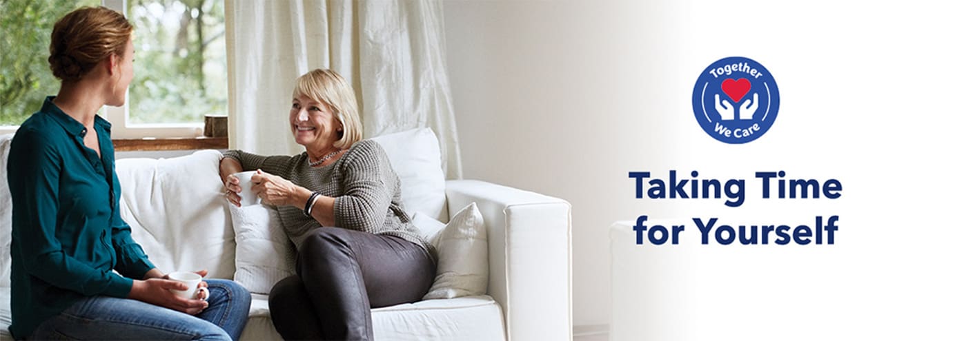 Mother and daughter, sitting on a white fabric couch and drinking from porcelain white coffee mugs, share smiles.